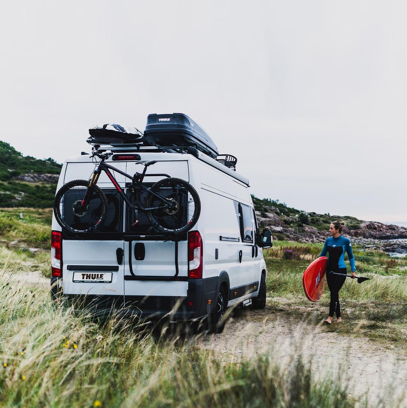 Campervan equipped with a Thule roof box and rear bike rack parked by the coast, as a surfer walks towards the sea with a board — representing van life adventure and outdoor travel gear.