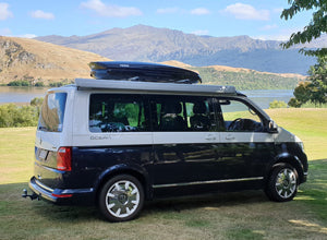VW campervan with a Thule roof box parked beside a mountain lake, showcasing van life travel and roof storage accessories.
