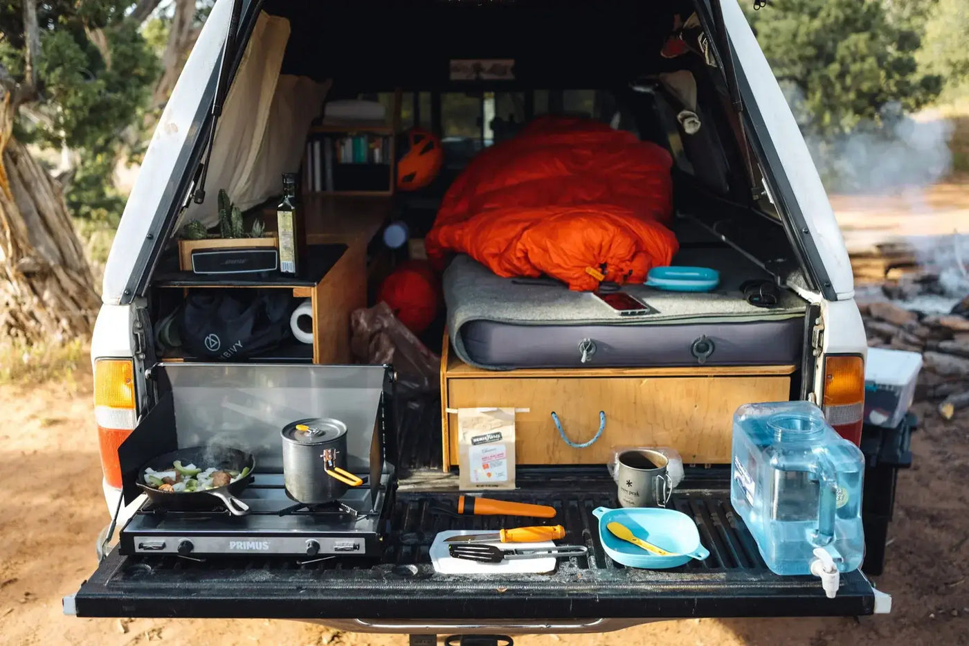 Rear view of a camper setup with a portable gas stove, cooking utensils, and sleeping area with a bright orange sleeping bag, showing efficient space use and off-grid van life living.