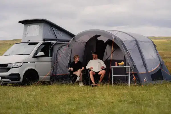 Couple relaxing outside a campervan fitted with a Vango Cove Air drive-away awning, showing spacious outdoor living area and easy-pitch camping setup on open grassland.