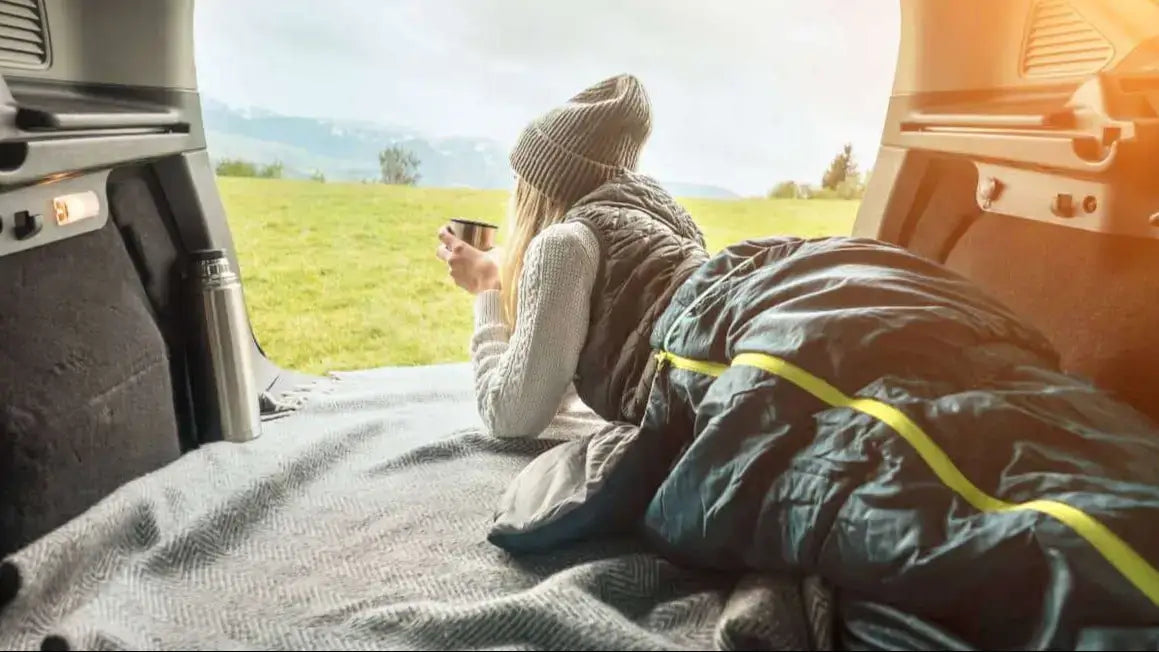 Woman relaxing in the back of a campervan with a sleeping bag and hot drink, looking out at a scenic morning view — representing cosy van life and minimalist travel comfort.