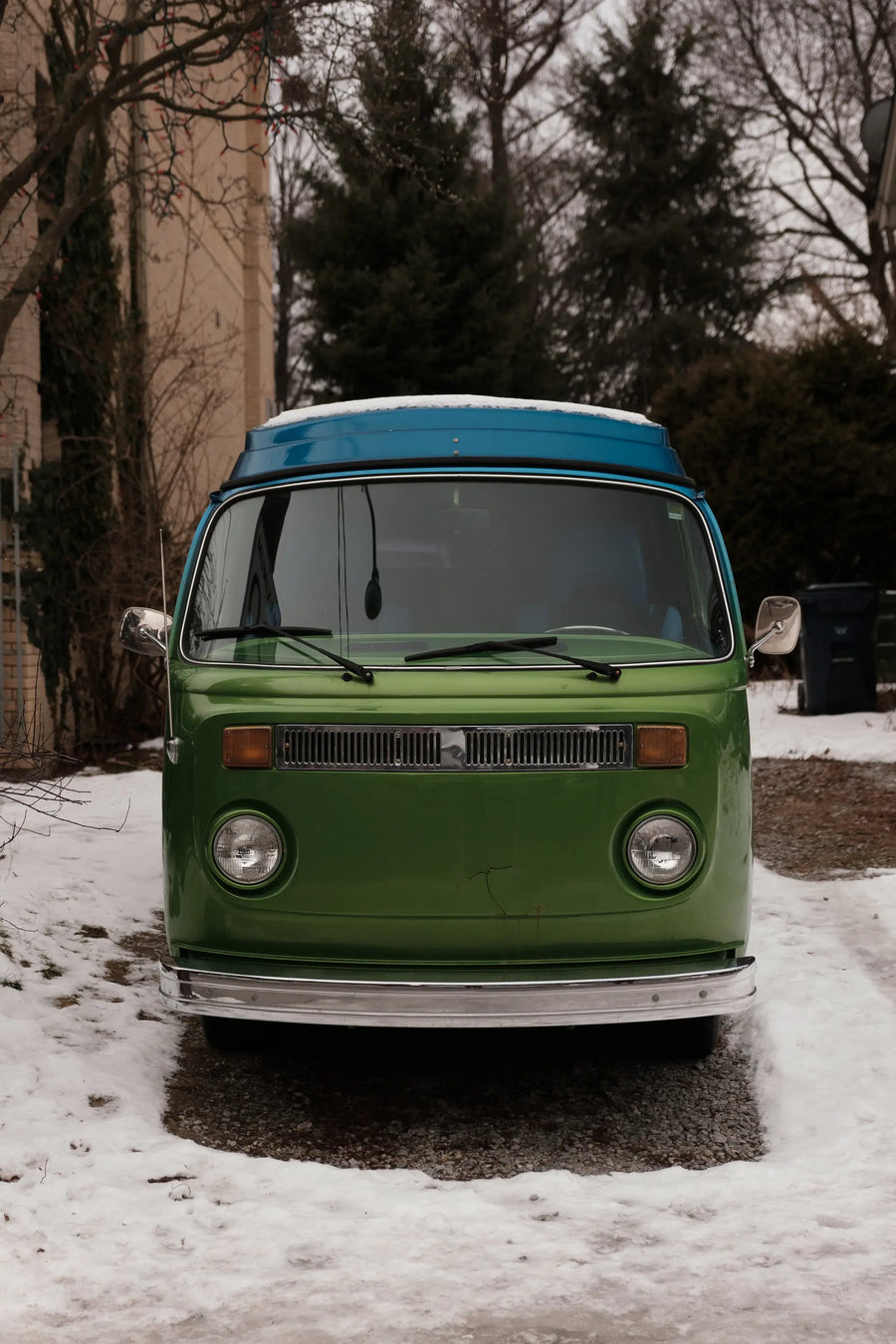 Front view of a classic green campervan with a blue pop-top roof parked on a snowy driveway, representing the van life travel lifestyle.