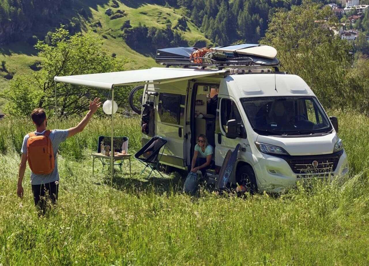 Motorhome parked in scenic countryside with awning extended and couple enjoying van life outdoors.